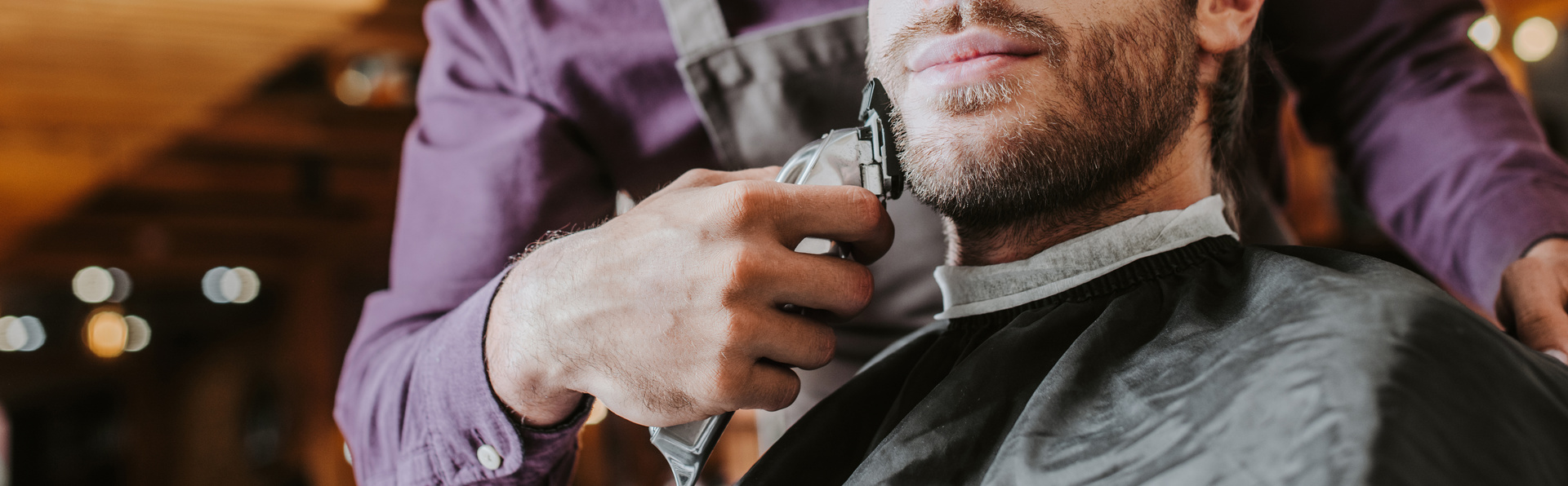 panoramic shot of barber holding trimmer while shaving bearded man
